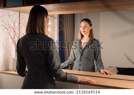 Self-confident Woman looking at her reflection into the mirror indoors. Beautiful interior design Royalty-Free Stock Photo #2118169514