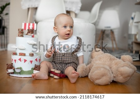 Portrait of infant little curious baby wearing square overall, celebrating happy birthday party. Presents and gifts with cake, decorated by bears cartoons. Big teddy bear near chair. Indoor apartment.