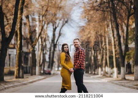 Stylish man in a red plaid shirt and a pregnant woman in a yellow dress are standing outdoors in nature holding hands. Pregnancy photography.