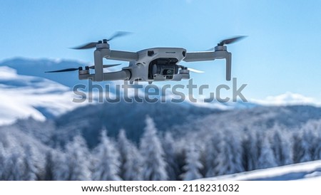 Drone with digital camera flying over winter landscape with snow covered mountains and trees. White remote controlled quadcopter hovering above snowy mountain peak. Close up shot of drone (UAV). Royalty-Free Stock Photo #2118231032