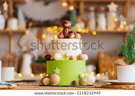 two-tiered white-green cake with balls, stars and a teddy bear on the kitchen table with electric garland for a children's holiday. the tradition of baking a birthday cake. professional confectioner.