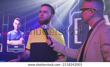 Adult man host with microphone asking questions to bearded gamer esportsman and announcing start of competition during gaming tournament