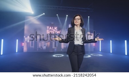 Happy presenter in a suit and glasses running onto the stage and announcing the start of the famous comedy, late-night show in an illuminated room with LED screen and 3D inscription Royalty-Free Stock Photo #2118242054