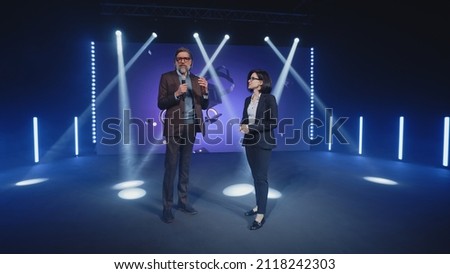 A female presenter interviewing a male developer of a new augmented reality device during the presentation of meta universe in a modern studio with spotlights, near the LED screen in 3D objects