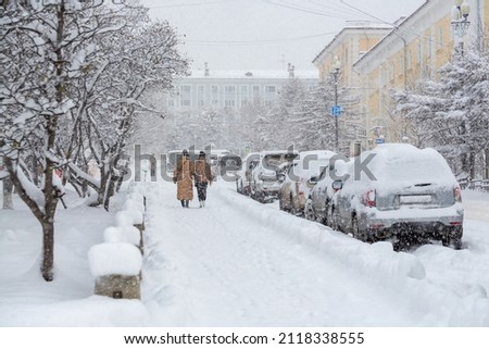 Snow covered city street during a heavy snowfall. Lots of snow on the sidewalk, cars and tree branches. Women walk around the winter city. Cold snowy weather. Magadan, Siberia, Russian Far East. Royalty-Free Stock Photo #2118338555