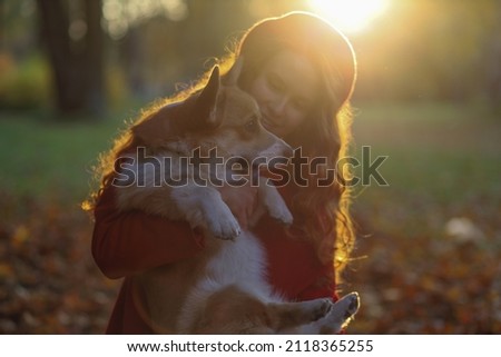 A girl with long red hair, in a red coat and beret gently hugs her corgi dog in an autumn park. Golden autumn, dog walking, friendship, sunset in the park, beautiful autumn picture