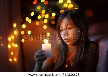 A portrait of a young beautiful girl holding a candle and looking at it with twinkling fairy lights as a background. Kids and Christmas.