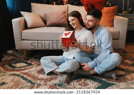 Young millennial man gives a gift to his beloved girlfriend on Valentine's Day. They sit on the floor in a cozy room. Congratulations and surprise on Valentine's Day.