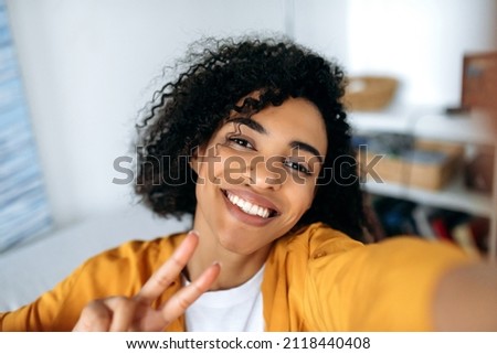 Cute beautiful joyful curly-haired African American girl, teenager, posing for selfie on smartphone, fooling around, having fun, looking at phone camera, smiling happily