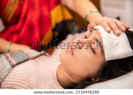 Indoor image of Indian matures mother sponging her ill daughter’s forehead to reduce the temperature down. She is having a high fever and lying in bed at home. She is wearing warm clothes. Royalty-Free Stock Photo #2118448280