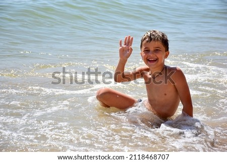 A white child splashes near the shore on a sunny sea beach, smiles and waves