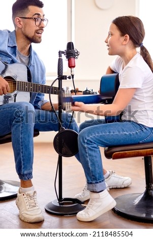 Handsome young man teaches little cute girl to play the guitar at home, singing in microphone. Happy Father's Day. Father and daughter in casual wear sit on couch in living room. I love you, dad.