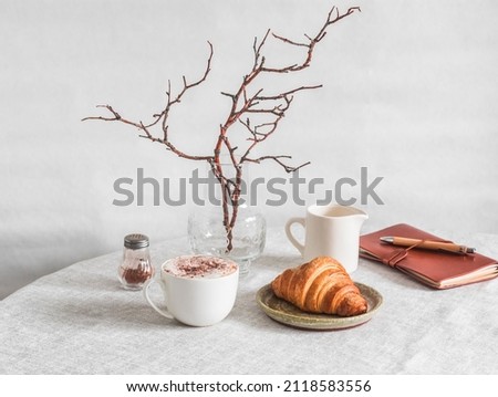 Cozy morning - cappuccino, croissant, vase with branches, leather planner notebook on the table in a bright cozy room. Cozy home interior      Royalty-Free Stock Photo #2118583556