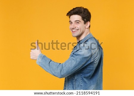 Side view of smiling young man guy in casual denim jacket posing isolated on yellow wall background studio portrait. People sincere emotions lifestyle concept. Mock up copy space. Showing thumb up Royalty-Free Stock Photo #2118594191