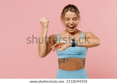 Young sporty athletic fitness trainer instructor woman wear blue tracksuit spend time in home gym use smart watch do winner gesture isolated on plain pink background. Workout sport motivation concept.