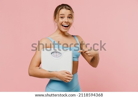 Young strong sporty athletic fitness trainer instructor woman wear blue tracksuit spend time in home gym point finger on scales isolated on pastel plain light pink background. Workout sport concept