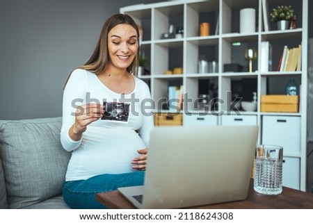 The selective focus of a cheerful pregnant woman who shows ultrasound photos while having a video call. She is happy for the child she is expecting, she shares happiness through video calls