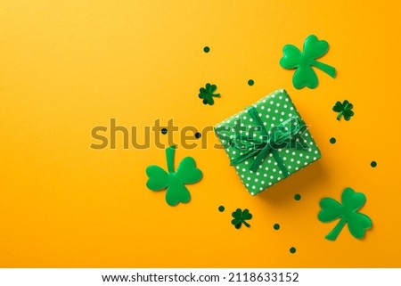 Top view photo of st patrick's day decorations green giftbox with polka dot pattern surrounded by shamrocks and clover shaped confetti on isolated yellow background with copyspace