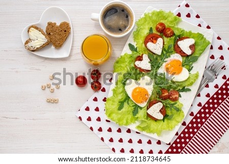 Breakfast on Valentines Day. A plate of fried heart-shaped eggs, cheese and tomatoes, coffee and juice, bread and butter on a white wooden table and the inscription I love you Breakfast for loved one