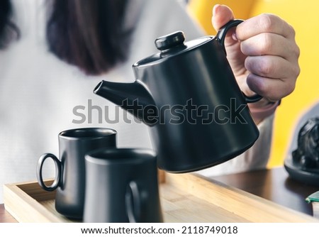 Close-up, a woman pours tea from a black teapot into black cups.