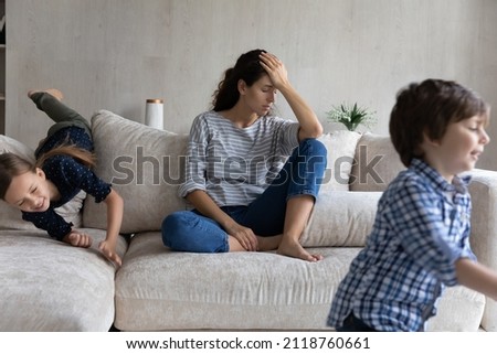 Tired young mother suffering from headache, feeling annoyed and exhausted by noisy kids running around, upset mom or babysitter sitting on couch, having problem with naughty active children Royalty-Free Stock Photo #2118760661