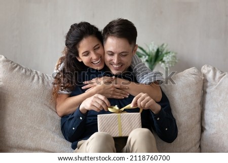 Loving girlfriend congratulating boyfriend, hugging from back, excited curious man holding gift box, sitting on couch, happy young couple celebrating anniversary or birthday at home together Royalty-Free Stock Photo #2118760700