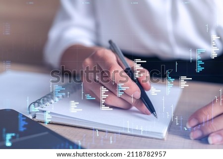A woman trader in formal wear writing down some ideas to research stock market to proceed right investment solutions. Wealth management concept. Hologram Forex chart over close up shot.