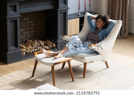 Full length mindful carefree young African American woman napping in comfortable armchair with legs on ottoman, breathing fresh air, sleeping in modern living room, enjoying peaceful weekend time. Royalty-Free Stock Photo #2118783791
