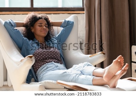 Full length millennial African American woman homeowner daydreaming in comfortable armchair with legs on footstool, breathing fresh air, sleeping resting, enjoying peaceful leisure time at home. Royalty-Free Stock Photo #2118783833