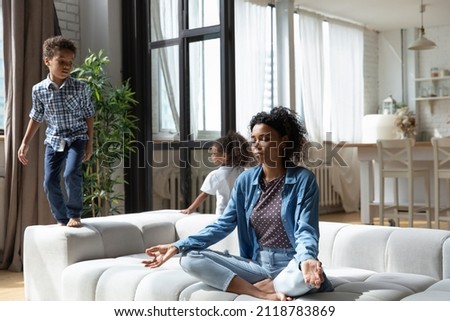 Happy mindful young African American mother sitting on comfortable couch in lotus position, relieving stress, doing yoga breathing exercises while joyful friendly children having fun at home. Royalty-Free Stock Photo #2118783869