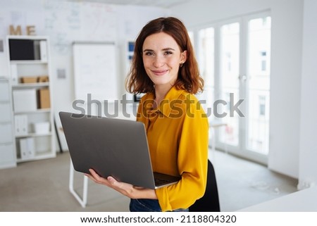 Smiling friendly young business manageress holding a laptop balanced on her arm looking at the camera as she poses in a high key open plan modern office Royalty-Free Stock Photo #2118804320
