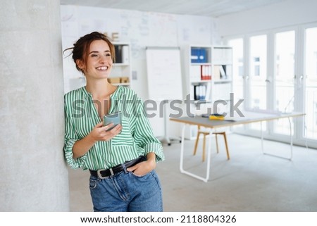 Happy young woman with a beaming vivacious smile leaning against an internal pillar in a spacious high key office looking to the side with copyspace Royalty-Free Stock Photo #2118804326