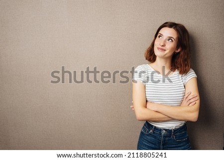 Thoughtful happy young woman smiling in anticipation as she stands with folded arms looking up to the side over a brown studio background with copyspace Royalty-Free Stock Photo #2118805241
