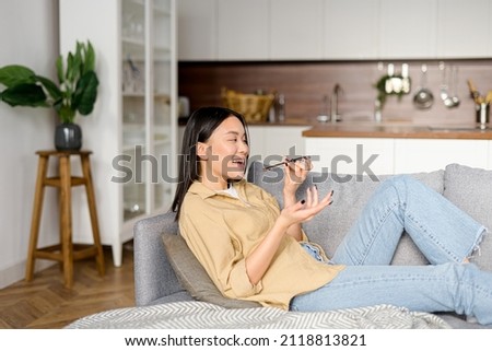 Charming young woman sitting on the sofa at home, recording and sending voice message. Female using voice recognition app on mobile phone, talking in the mic of the smartphone