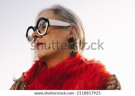 Pensive senior woman looking away while standing outdoors. Stylish elderly woman wearing colourful casual clothing and eyeglasses. Mature woman reflecting on memories of the past. Royalty-Free Stock Photo #2118817925