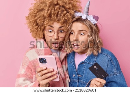 Photo of two shocked beautiful women stare at smartphone hold credit card dont expect to get bonus and cashback make online payment dressed in fashionable clothes isolated over pink background