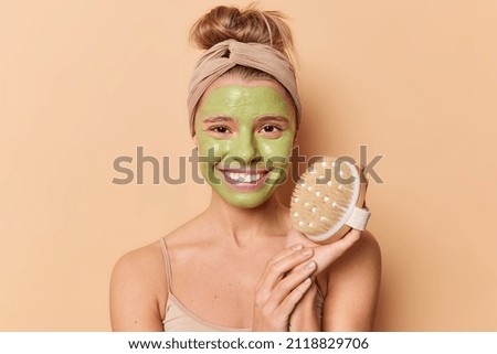 Horizontal shot of glad young European woman takes care of face applies green nourishing mask holds body brush for making massage has combed hair wears headband isolated over brown background