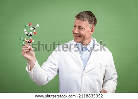 Scientist with white coat holds model of molecules in hand and stands in front of green background