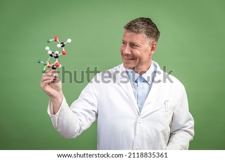 Scientist with white coat holds model of molecules in hand and stands in front of green background