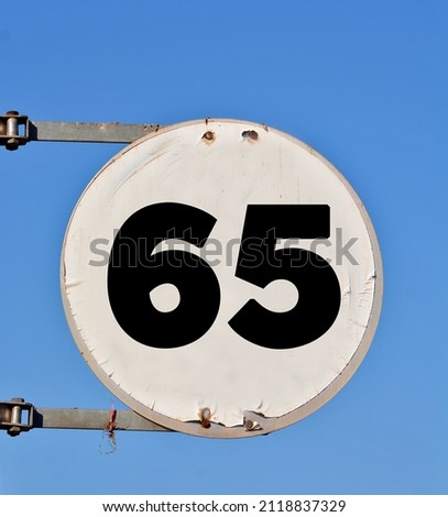 A round white sign, showing a black coloured number sixty five (number 65) 