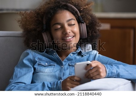 Smiling peaceful young gen Z teen Black girl wearing big wireless headphones, using smartphone, enjoying relaxing romantic music resting on comfortable couch at home with closed eyes Royalty-Free Stock Photo #2118840440