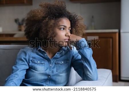 Frustrated pensive teenage African girl sitting on couch at home, looking away, thinking over heartbreak problems, offence, feeling bored, suffering from depression. Youth problems concept Royalty-Free Stock Photo #2118840575
