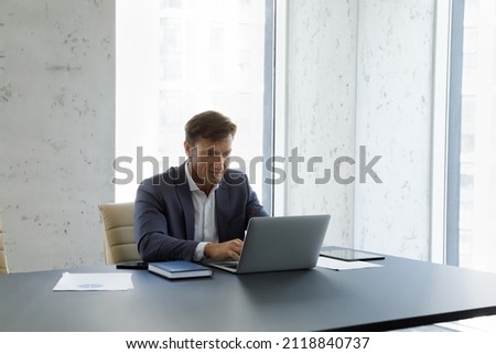 Busy confident mature business leader working at computer in office. Male boss sitting at workplace, typing on laptop, surfing, browsing internet, making video call, using online professional app