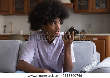 Serious teen African hipster guy recording audio message on mobile phone, using voice recognition app, making cellphone call, talking to virtual assistant service, sitting on home sofa