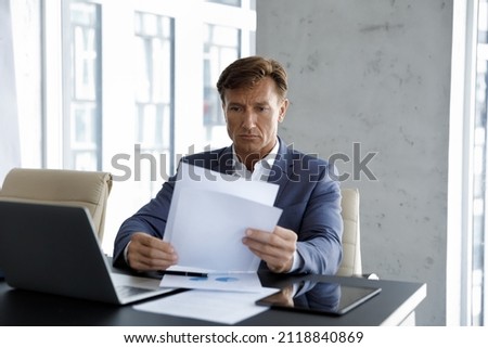 Focused mature company executive reviewing sales reports, checking documents at laptop on workplace, reading corporate papers, analyzing marketing data, statistic research. Business, paperwork concept Royalty-Free Stock Photo #2118840869