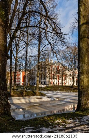 Vertical photo of Jelgava Palace framed with leafless tress and frozen path reflecting building at sunny clear winter day. Historical building in the city park at winter day.