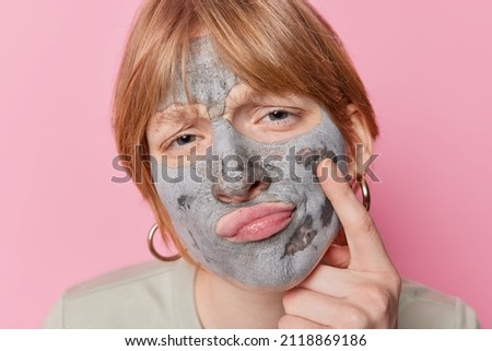 Headshot of redhead woman looks attentively examines skin applies nourishing clay mask for rejuvenation keeps finger on cheek wears earrings isolated over pink background. Beauty and wellness