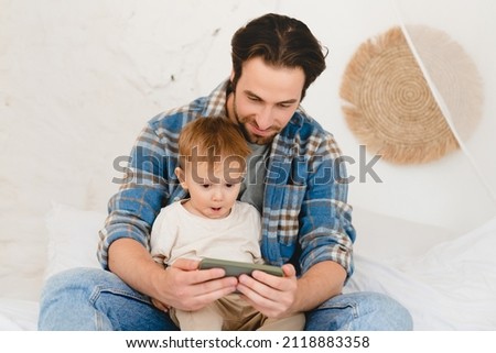 Young father dad spending time with daughter newborn infant child son toddler kid at home, watching cartoons on smart phone online at home. Fatherhood concept.