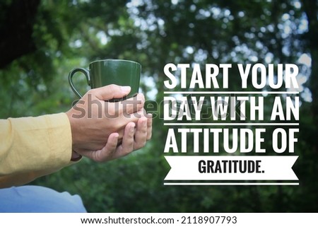 Inspirational quote - Start your day with an attitude of gratitude. With person holding a cup of tea or coffee in hands on green nature park background. Morning grateful thankful concept