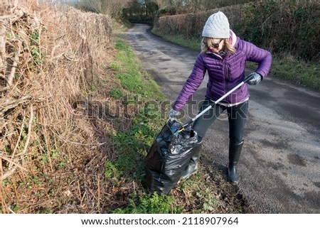 A lady uses a litter pick up tool to collect a plastic bottle and place it in a black bin bag.She wears plastic gloves and stands on verge of a rural country lane.Pollution.Garbage.
 Royalty-Free Stock Photo #2118907964